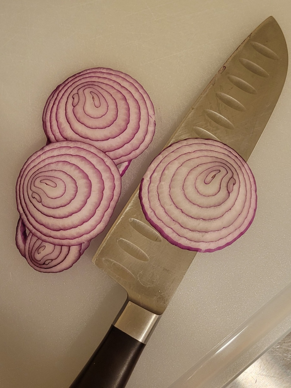 Thin slices of red onion and a silver chef's knife with a brown handle lay on a white cutting board