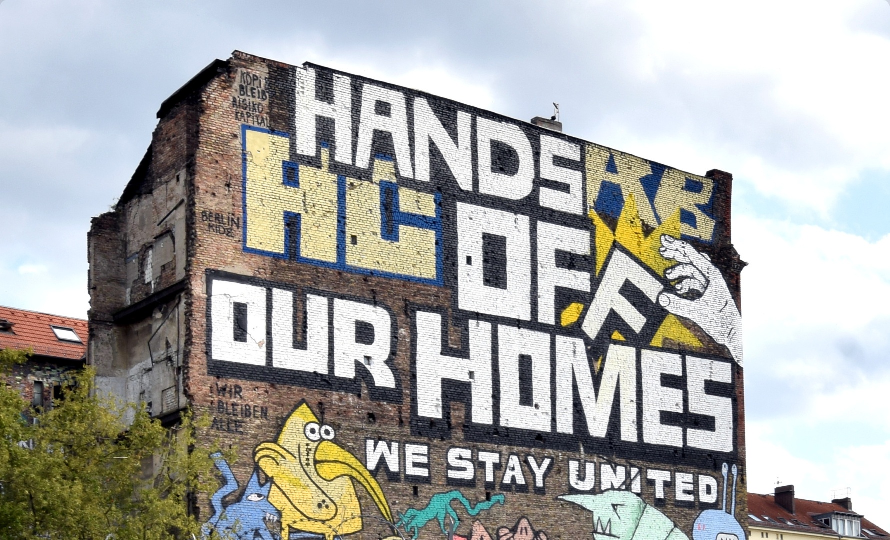 Foto eines großen Graffitis über die ganze Wand eines mehrstöckigen Hauses. Text: Hands off our homes; Köpi bleibt Risikokapital; Wir bleiben alle; Stay United. Darunter sind noch Bilder eines gelben Vogels zu sehen. 
Eine Hand greift nach dem F in dem Wort off.