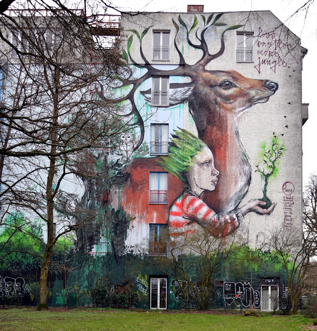 Mural an einer  Hauswand. Zu sehen sind ein großere Hirsch und daneben ein Person mit grünen Blättern als Haare und einem weiß-rot gestreiften Pulli. In der rechten Hand hält die Person einen kleinen Baum um den Vögel fliegen. Am rechten oberen Rand steht: "Less castles more jungle". Am rechten unteren Rand ist die Signatur zu finden: "Hera of Herakut + juli Jah + friends. For the green forest fund. 2023"