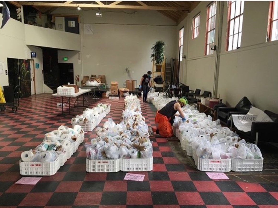 Photo of a large open room, with red and black tile floors, white walls, and high ceilings. On the floor there are three rows of grocery bags filled with essential items and food. There are many bags of groceries, and two people wearing masks and gloves organizing the rows of bags.