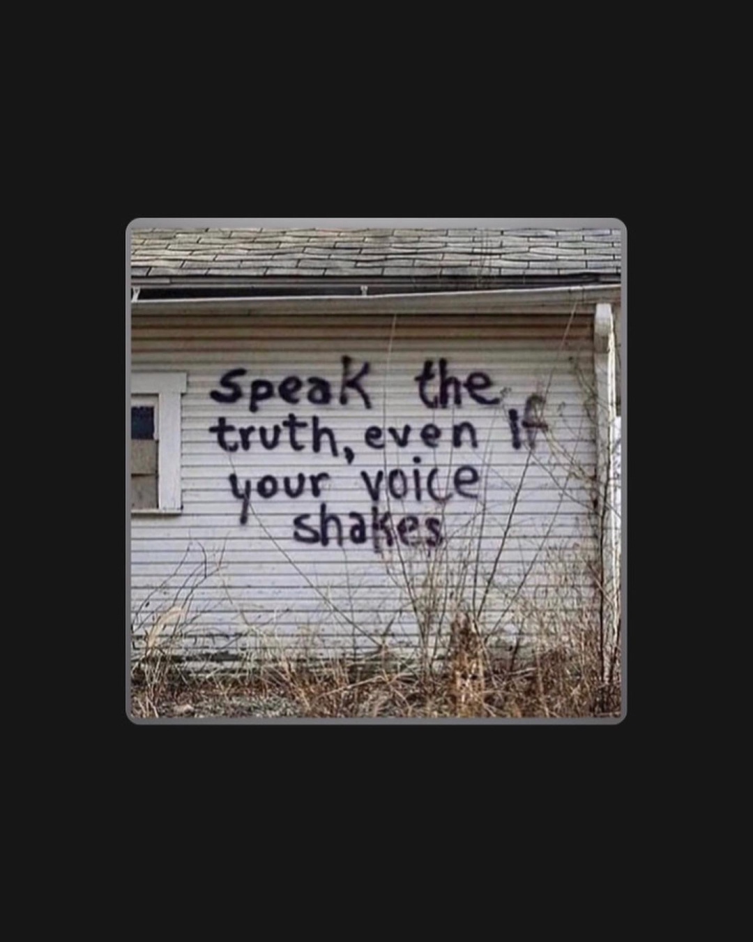 A photo of the paneling wall on the side of a dingy, old, white house, with a boarded up window, and black spray paint writing that reads: “speak the truth, even if your voice shakes”