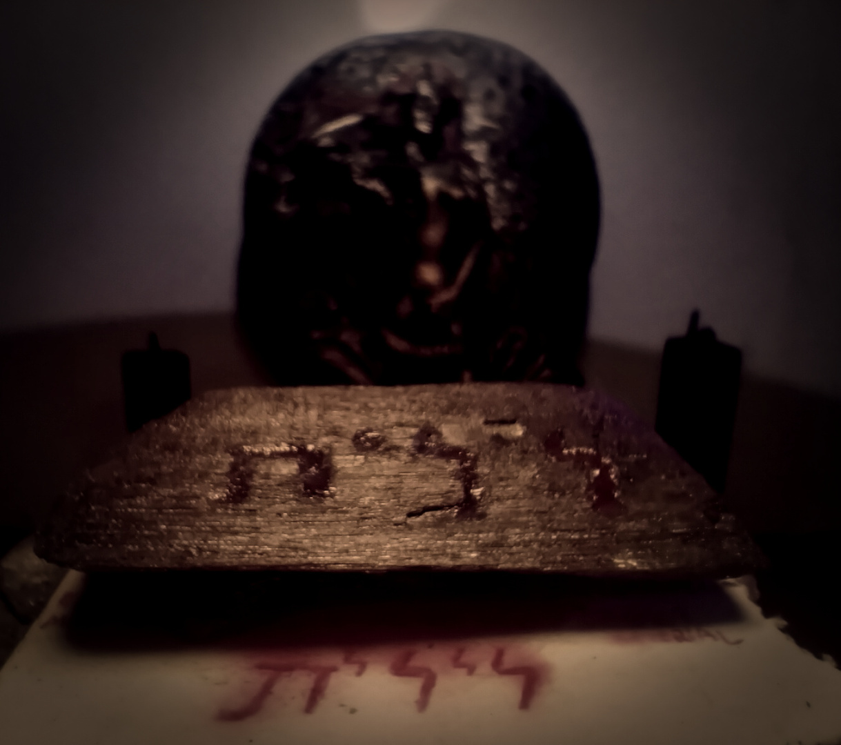 A highly symmetrical photo with vignette and chiaroscuro effects, the latter of which is the result of the setup involving two warm white light sources and one UV flashlight. There are few post-edition effects aside from the vignette. The scene depicts a very close (4 cm from the camera) piece of charcoal, to which the camera is focused using macro mode (so all the other items from the scene are blurry). The piece of charcoal is visibly thin with razor-sharp edges, and there's an engraving on top of it, which reads the sequence of hebrew letters Lamedh-yodh-lamedh-yodh-taw (L-Y-L-Y-Th) from right to left. The engraving is filled with red nail polish. Under the charcoal, there's a piece of paper with just the bottom part of it visible. The bottom part of the paper contains the exact same Hebrew inscription, also in red. Behind, there's a bronze statue depicting Aphrodite, flanked by two unlit black candles. The gray wall behind reflects a circular light spot above Aphrodite's head.